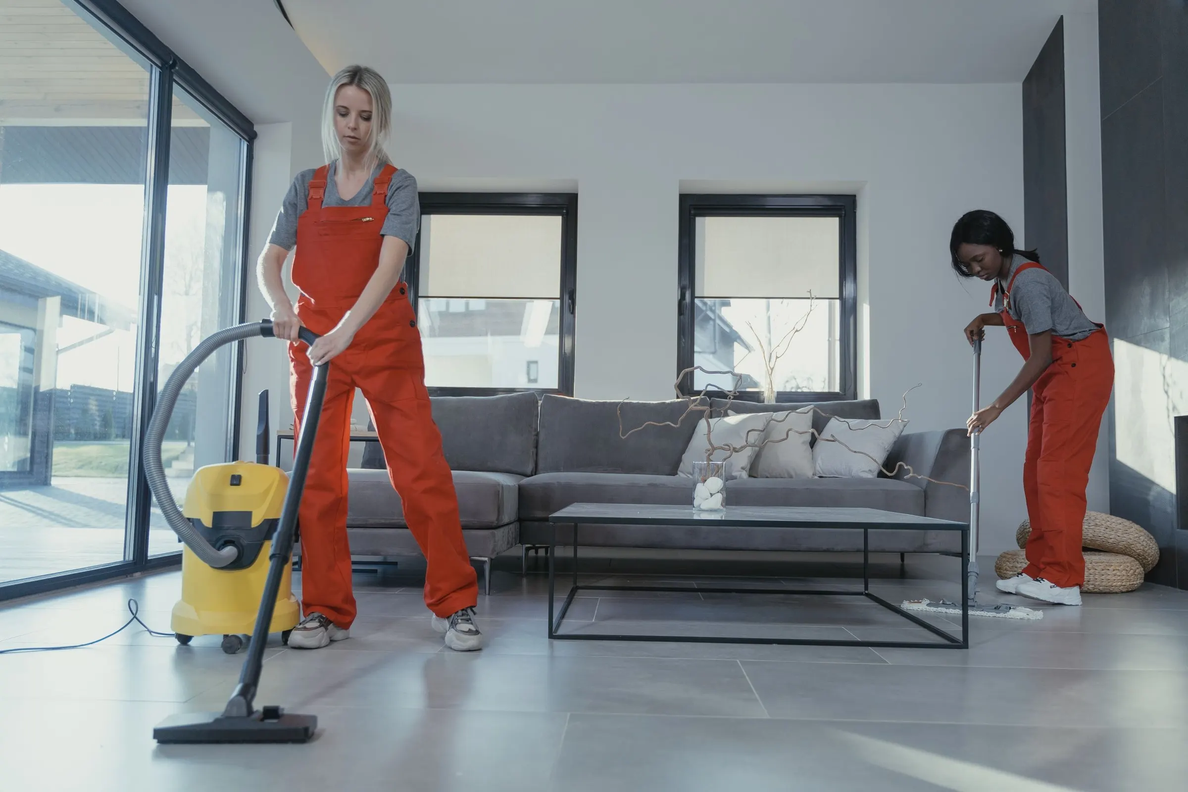 Two cleaners in red overalls working through a living room with a vacuum and a mop