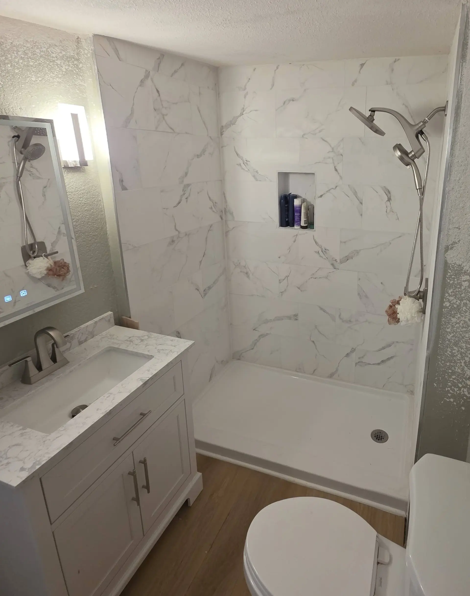 A bathroom with a marble-look tile shower on a white base, a white vanity and a plank floor