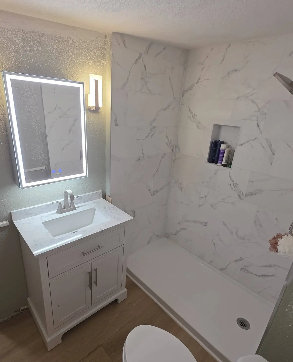 A remodeled bathroom with a marble-look tile shower, a white vanity under a lit mirror and a plank floor