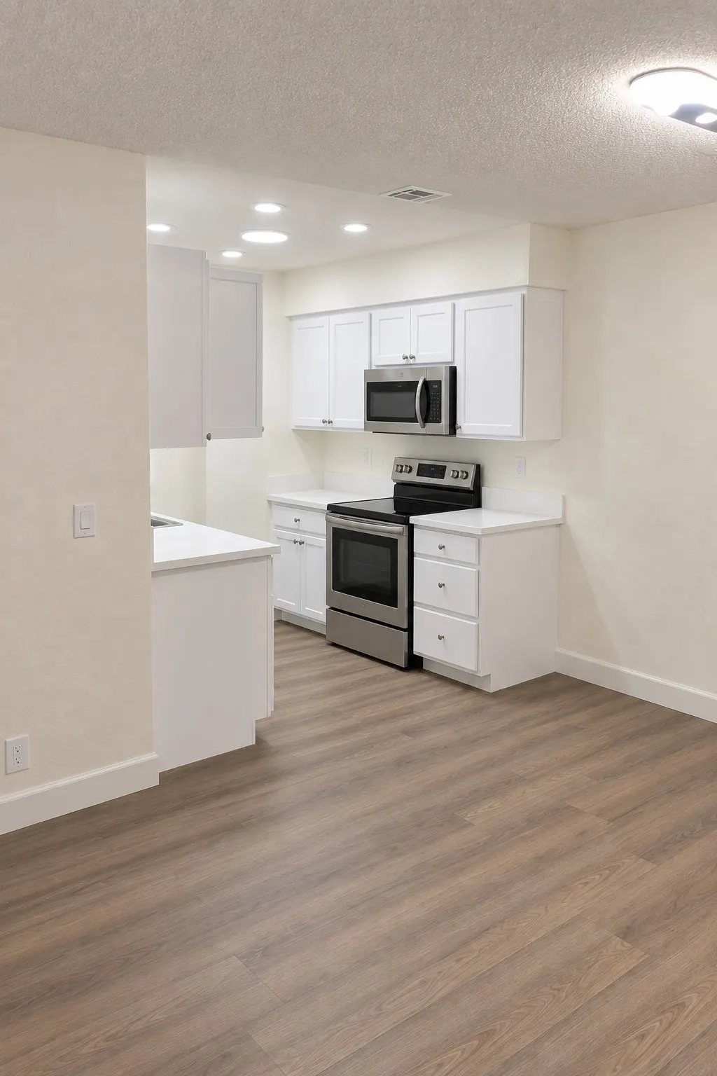 An empty living room with a wood-look plank floor and a white kitchen with a stainless range at the back